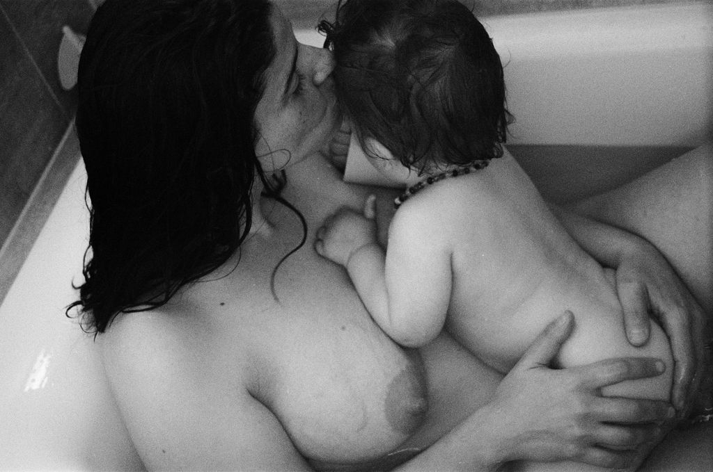 A black and white film photograph of a postpartum mother with dark hair in the bath of a Launceston home with her infant. His face is turned away as she kisses the top of his head.